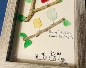 May include: Framed artwork featuring three colorful glass birds perched on branches with green glass leaves. The artwork includes the handwritten text "Every little thing Gonna be alright" and a line of sketched flowers. The frame is gray.
