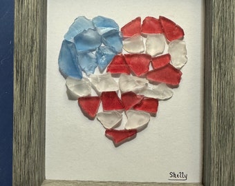 Op de afbeelding: Een ingelijst hartvormig kunstwerk gemaakt van rood, wit en blauw zeeglas. Het kunstwerk is gesigneerd met "Shelly".