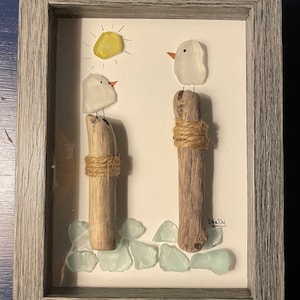 May include: A framed artwork featuring two white glass birds perched on driftwood posts, with a yellow sun and blue glass sea glass pieces at the bottom. The artwork is signed "Shelly".