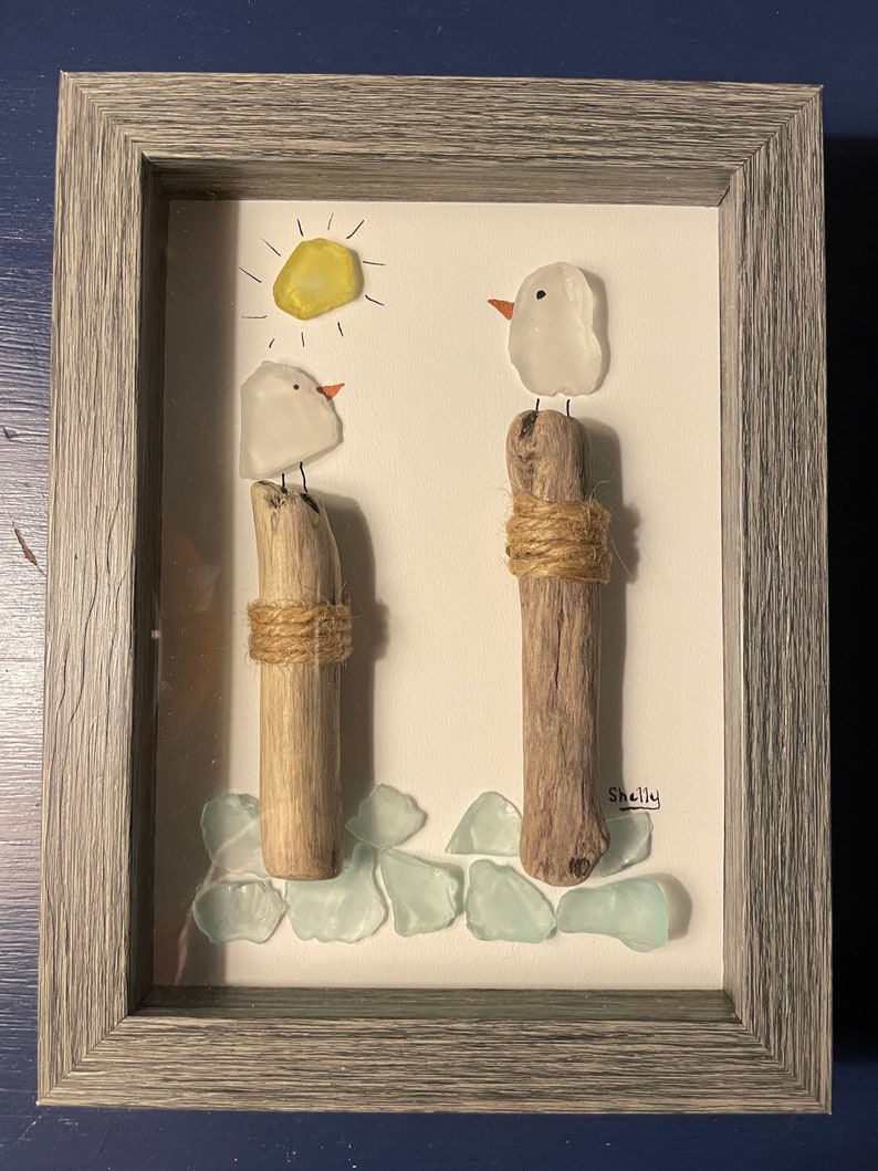 May include: A framed artwork featuring two white glass birds perched on driftwood posts, with a yellow sun and blue glass sea glass pieces at the bottom. The artwork is signed "Shelly".