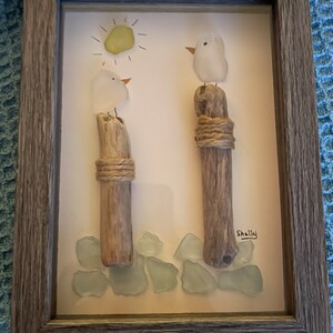 May include: A framed artwork featuring two white glass birds perched on driftwood posts, surrounded by light blue sea glass. The artwork includes a yellow sun with rays and the signature "Shelly".