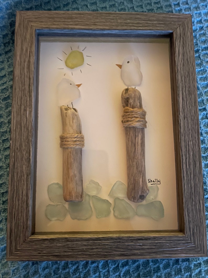 May include: A framed artwork featuring two white glass birds perched on driftwood posts, surrounded by light blue sea glass. The artwork includes a yellow sun with rays and the signature "Shelly".