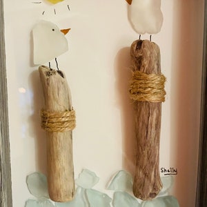 May include: A framed artwork featuring two white glass birds perched on driftwood posts, with a yellow glass sun and blue glass pieces at the bottom. The artwork is signed "Shelly".