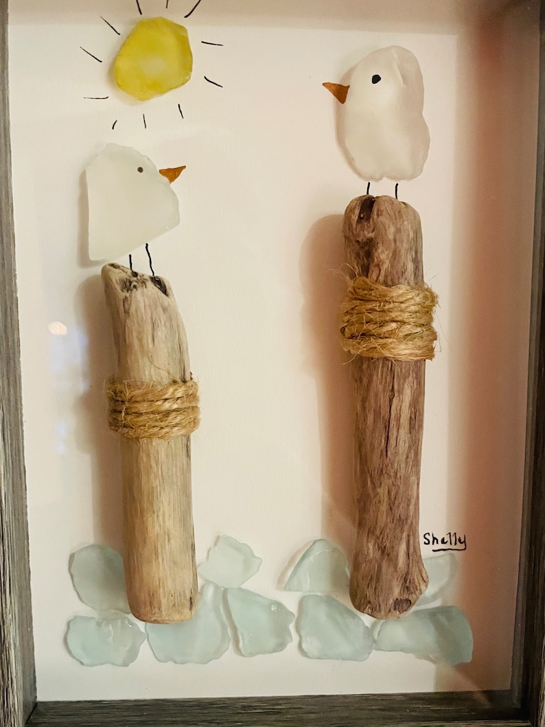 May include: A framed artwork featuring two white glass birds perched on driftwood posts, with a yellow glass sun and blue glass pieces at the bottom. The artwork is signed "Shelly".