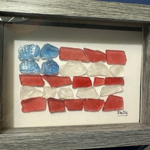 Könnte beinhalten: Gerahmtes Kunstwerk, das die amerikanische Flagge darstellt, hergestellt aus rotem, weißem und blauem Meeresglas. Das Design der Flagge befindet sich vor einem weißen Hintergrund in einem grauen Holzrahmen. Die Signatur des Künstlers, "Shelly", ist sichtbar.