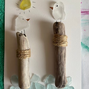 May include: Two white glass birds perched on driftwood posts with twine wrapped around the base. The birds are facing different directions. A yellow sun with rays is drawn above the birds. The background is white with light blue glass pieces scattered around the base of the posts.
