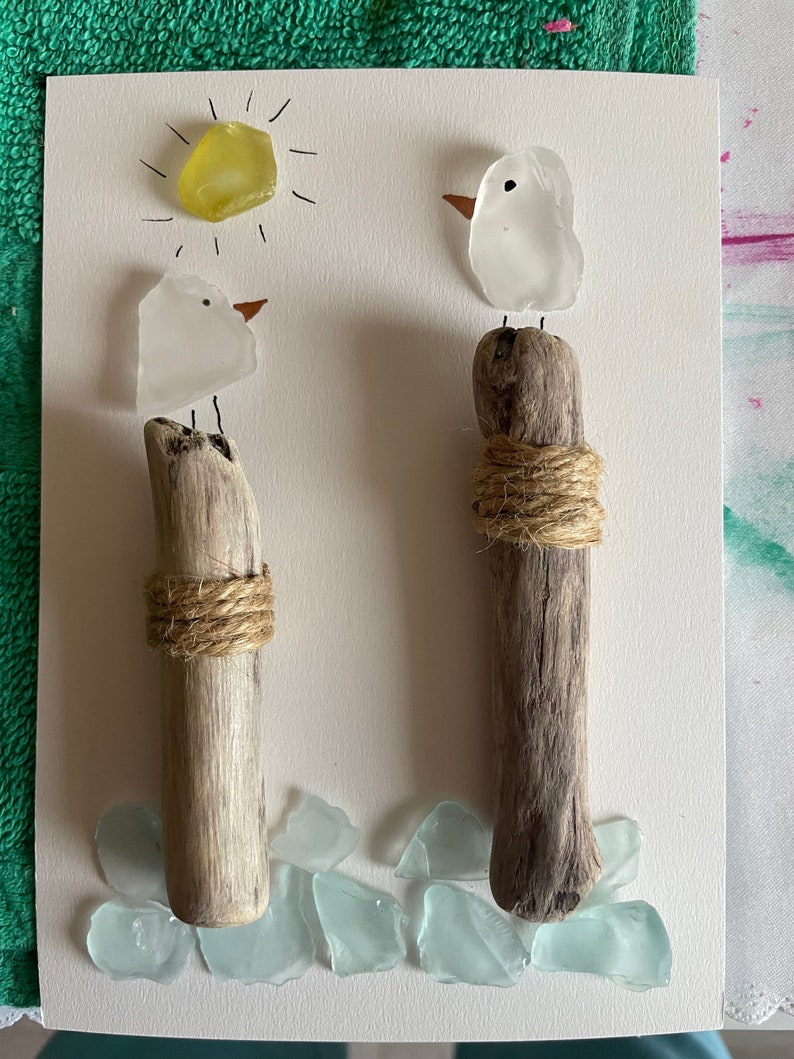 May include: Two white glass birds perched on driftwood posts with twine wrapped around the base. The birds are facing different directions. A yellow sun with rays is drawn above the birds. The background is white with light blue glass pieces scattered around the base of the posts.