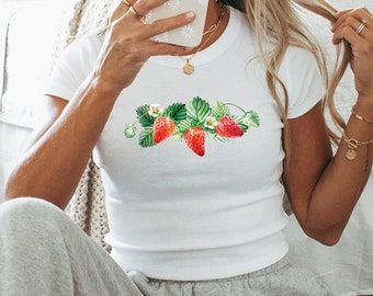 Op de afbeelding: Een vrouw draagt een wit T-shirt met een aardbeienprint en grijze sweatpants. Ze zit op een tapijt met een patroon en houdt een witte telefoon voor haar gezicht.