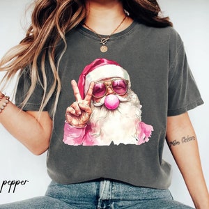 May include: A gray t-shirt with a graphic of Santa Claus wearing pink sunglasses and blowing a bubble. Santa is giving the peace sign with his left hand. The text "pepper" is visible in the bottom left corner of the image.