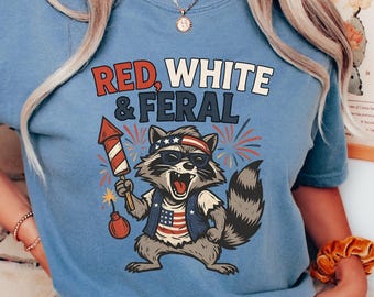 May include: A blue t-shirt with the text "RED, WHITE & FERAL" and a raccoon graphic wearing sunglasses and a patriotic vest, holding a firework and a bomb. The shirt is paired with white pants.