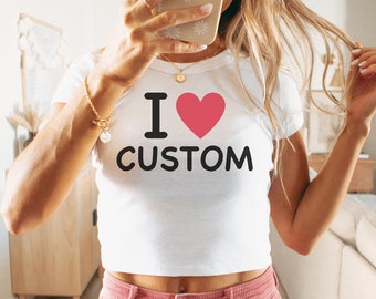 Op de afbeelding: Een wit gecropped t-shirt met een zwarte grafische afbeelding die "Ik hou van custom" zegt met een roze hart. Het model draagt een roze corduroy broek.
