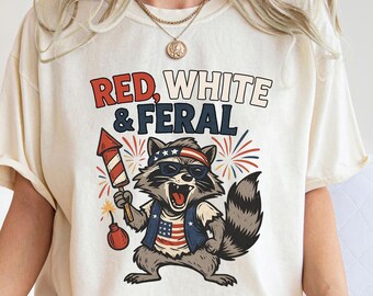 May include: Ivory-colored t-shirt featuring a raccoon wearing sunglasses, a patriotic vest, and a headband. The shirt has the text "Red, White & Feral" and a firework graphic. The raccoon is holding a firework.