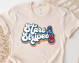 Può includere: Una maglietta beige con una grafica in stile retrò che dice "Stars & Stripes" con un segno di pace al centro. Il segno di pace è decorato con stelle rosse, bianche e blu.
