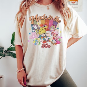 May include: Cream-colored t-shirt with a retro graphic that reads "Made in the 80s." The graphic features cartoon characters and a rainbow design. The shirt is a short-sleeved, casual style.
