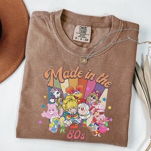 May include: A brown t-shirt with the text "Made in the 80s" and a graphic of cartoon characters. The shirt has a crew neck and a small tag at the neckline. A silver necklace with a pendant is draped across the shirt.