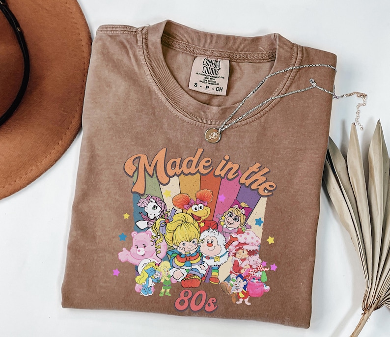 May include: A brown t-shirt with the text "Made in the 80s" and a graphic of cartoon characters. The shirt has a crew neck and a small tag at the neckline. A silver necklace with a pendant is draped across the shirt.