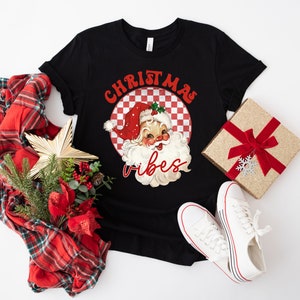 May include: Black t-shirt with a retro Santa Claus graphic and the words "Christmas Vibes" in red. The image includes a red and green plaid scarf, a gift box with a red bow, and white sneakers.