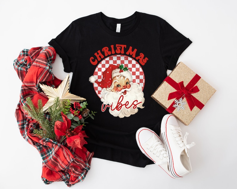 May include: Black t-shirt with a retro Santa Claus graphic and the words "Christmas Vibes" in red. The image includes a red and green plaid scarf, a gift box with a red bow, and white sneakers.