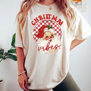 May include: Cream-colored t-shirt featuring a retro Santa Claus graphic with the words "Christmas Vibes" in red. The Santa illustration is set within a red and white checkered circle, evoking a vintage holiday aesthetic.