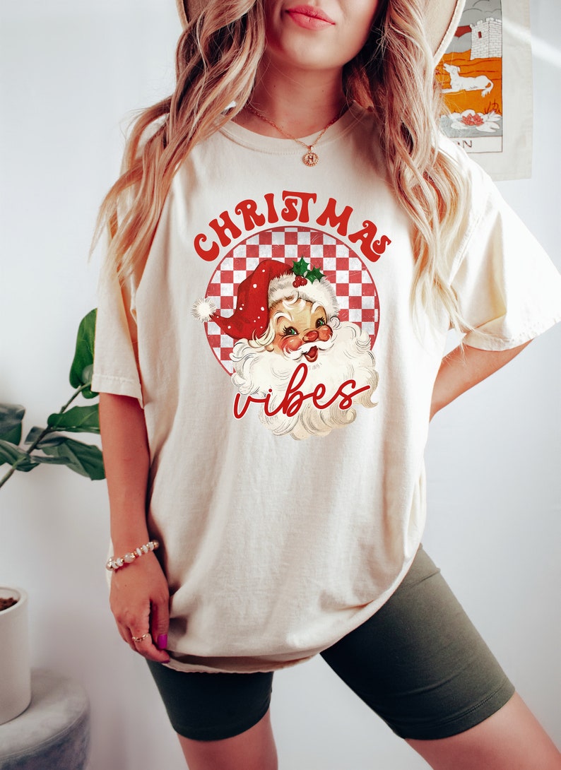 May include: Cream-colored t-shirt featuring a retro Santa Claus graphic with the words "Christmas Vibes" in red. The Santa illustration is set within a red and white checkered circle, evoking a vintage holiday aesthetic.