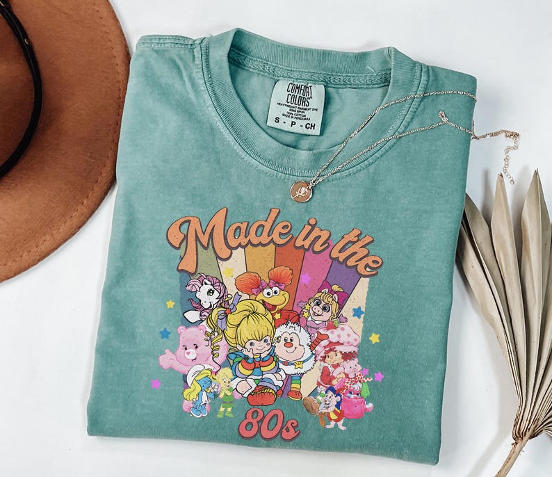 May include: A teal Comfort Colors t-shirt with a graphic that says "Made in the 80s" with cartoon characters. A gold necklace with a pendant is on the shirt. The shirt is folded on a white surface.