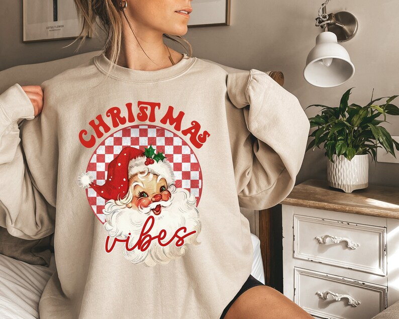 May include: Beige sweatshirt featuring a retro Santa Claus graphic with the words "CHRISTMAS" and "vibes" in red. The Santa graphic is inside a red and white checkered circle, evoking a vintage holiday theme.