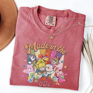 May include: A red t-shirt with a retro graphic that says "Made in the 80s" with cartoon characters. The shirt has a crew neck and short sleeves. A gold necklace with a pendant is on the shirt.