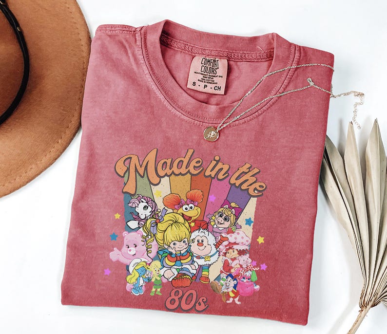 May include: A red t-shirt with a retro graphic that says "Made in the 80s" with cartoon characters. The shirt has a crew neck and short sleeves. A gold necklace with a pendant is on the shirt.
