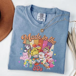 May include: A blue Comfort Colors t-shirt with a graphic that says "Made in the 80s." The graphic features cartoon characters and a rainbow design. A gold necklace is draped across the shirt.