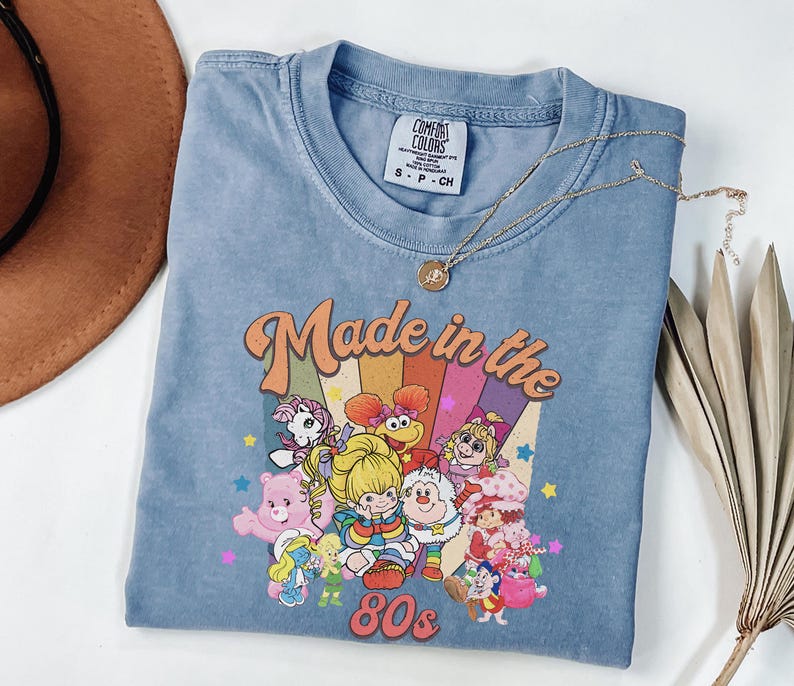 May include: A blue Comfort Colors t-shirt with a graphic that says "Made in the 80s." The graphic features cartoon characters and a rainbow design. A gold necklace is draped across the shirt.