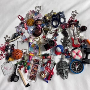 May include: A collection of keychains with various charms, including a basketball, a camera, a guitar, a dumbbell, and a compass. The keychains are adorned with beads and other decorative elements. Some keychains have text, such as "Movie Friday?"