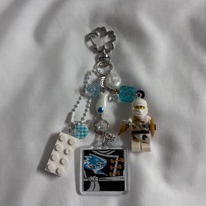 May include: A handmade keychain featuring a Lego minifigure, white Lego brick, and a clear square charm with a graphic design. The keychain includes blue and white beads, a silver-tone clasp, and a small chain.