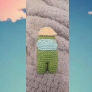 May include: A green crocheted plush toy shaped like a character from the game Among Us. The toy is wearing a yellow hat and a light blue shirt.