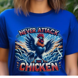 Puede incluir: Una camiseta azul con un gráfico de un gallo con un diseño azul y rojo. El gallo está de pie sobre una nube con rayos. El texto "Never Attack The Chicken" está escrito en letras rojas y blancas.