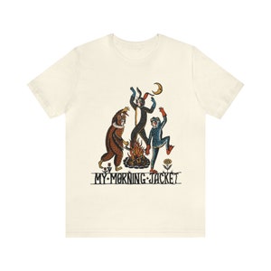 May include: A white t-shirt featuring a graphic print of three figures dancing around a bonfire. The figures are a brown bear, a black rabbit, and a blue frog. The text "MY MORNING JACKET" is printed below the figures.
