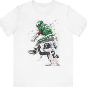 May include: A white t-shirt with a green and black watercolor illustration of two football players in action. The player in the green jersey is leaping over the player in the black jersey. The player in the green jersey has the number 22 on his jersey. The player in the black jersey has the number 22 on his jersey.