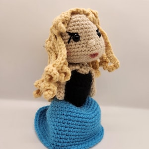 May include: A crocheted doll with blonde hair, a black dress, and a blue skirt. The doll has a crocheted face with black eyes and a pink mouth.