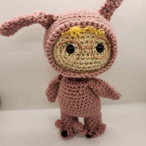 May include: A handmade crochet doll in a pink bunny costume with a light beige face and yellow hair peeking out. The doll has wire-rimmed glasses and black eyes. The bunny ears are upright. The doll's feet have black paw pads.