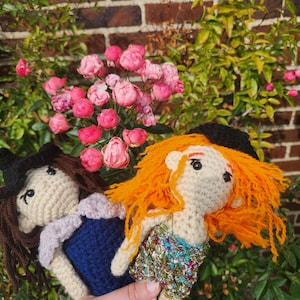 May include: Two handmade crochet dolls featuring yarn hair and detailed outfits. One doll has brown hair, a black hat, and a blue dress with a light purple collar. The other doll has bright orange hair, a black hat, and a multi-colored sequined top.