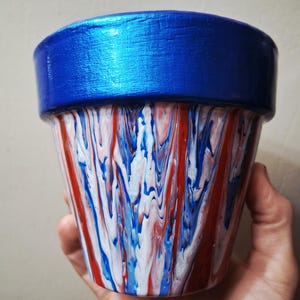 Op de afbeelding: Een taps toelopende plantenpot met een effen metallic blauwe rand en een lichaam versierd met een gemarmerd verfpatroon in rood, wit en blauw. Het ontwerp bestaat uit verticale, vloeiende kleurstrepen die over het oppervlak in elkaar overlopen.