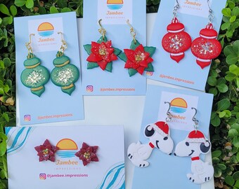 Puede incluir: Cinco tarjetas con aretes de temática navideña. Los aretes están hechos de arcilla polimérica y presentan una variedad de diseños, incluyendo poinsettias, adornos y Snoopy. Las tarjetas son blancas con acentos azules y verdes y presentan el texto "Jambee Impressions" y el nombre de usuario de las redes sociales "@jambee.impressions".