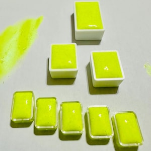 May include: A collection of neon yellow watercolor paint pans in various arrangements. Some are in white plastic containers, while others are in clear plastic containers. A streak of paint is visible on the left.