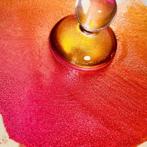 May include: A clear glass bottle stopper with a spherical top and a gold-coloured base. The stopper sits on a surface with a gradient of red and orange hues, creating a vibrant display.