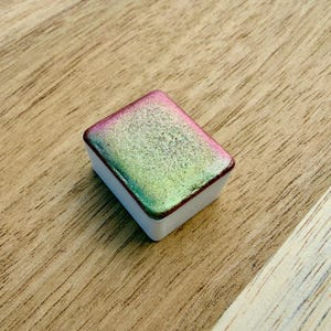 Puede incluir: Una pastilla de acuarela rectangular con base blanca y superficie brillante. La pintura tiene un degradado de colores, incluyendo rosa, verde y dorado. La pastilla está sobre una superficie de madera.