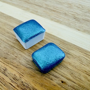 Könnte beinhalten: Zwei quadratische, blaue Aquarellfarben mit glitzerndem Finish. Die Farbtöpfchen stehen auf einer Holzoberfläche mit einem hellen Streifen. Die Farbtöpfchen haben eine weiße Basis.