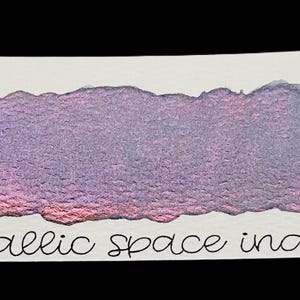 Puede incluir: Una muestra de pintura de acuarela índigo espacial metálico sobre papel blanco. La pintura es una mezcla de tonos morados, rosas y azules, con un acabado brillante y metálico. El texto "no. 63" y "metallic space indigo" están escritos en el papel.