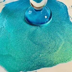 Op de afbeelding: Een close-up van een helder glazen object met een afgeronde basis, rustend op een glinsterend turquoise en blauwgroen oppervlak. Het oppervlak heeft een textuur, korrelig uiterlijk, met hints van groen en wit.