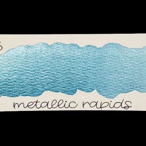 Könnte beinhalten: Ein Farbmuster aus metallisch blauer Aquarellfarbe auf einer weißen rechteckigen Karte. Die Karte trägt oben den handgeschriebenen Text "no. 128" und unten "metallic rapids". Die Farbe hat ein strukturiertes, schimmerndes Aussehen.