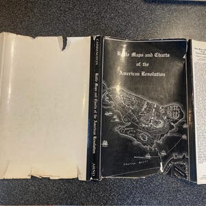 May include: An open book titled "Battle Maps and Charts of the American Revolution." The cover displays a detailed black and white map. The spine of the book reads "Carrington" and "Arno."