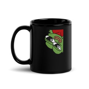 Puede incluir: Taza de cerámica negra con una rana de dibujos animados verde y roja asomándose por el lado. La rana lleva un sombrero rojo y tiene ojos grandes.